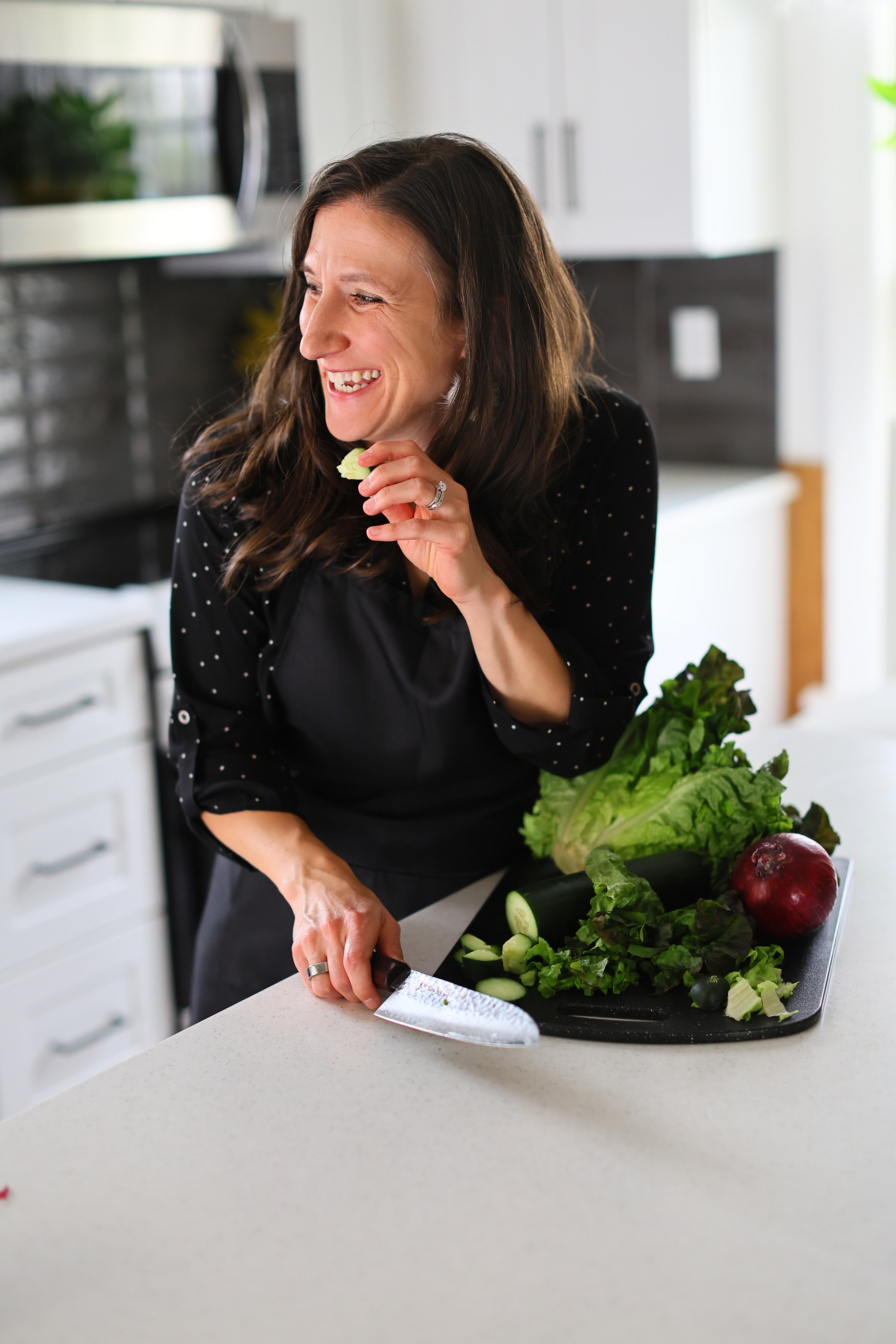 Dr. Anne Bodensteiner in black apron slicing cucumber on kitchen counter with lettuce and red onion nearby.