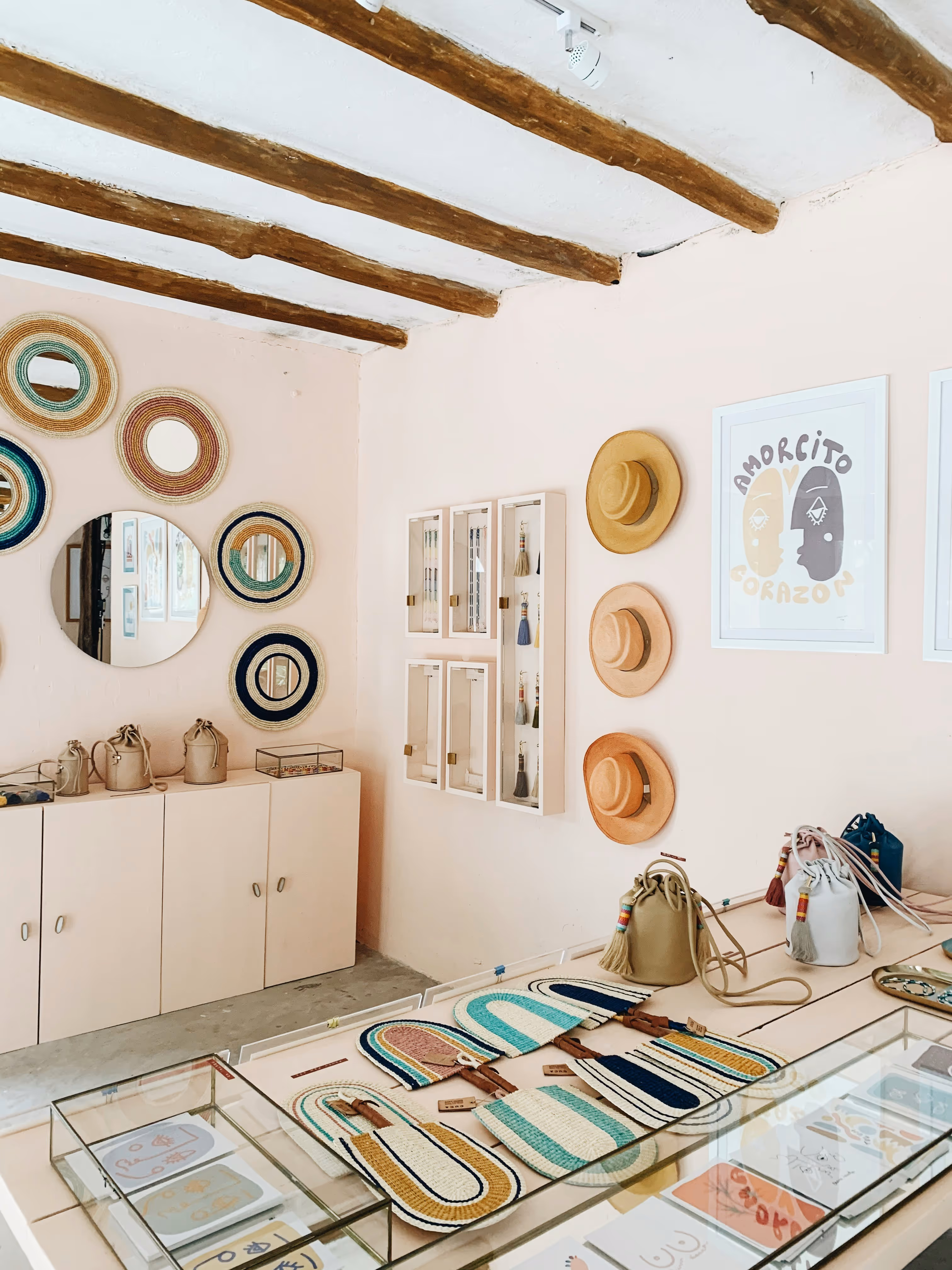 Bright boutique interior with wooden ceiling beams, hats on the wall, woven bags on a display table, and circular woven wall decor around a round mirror.