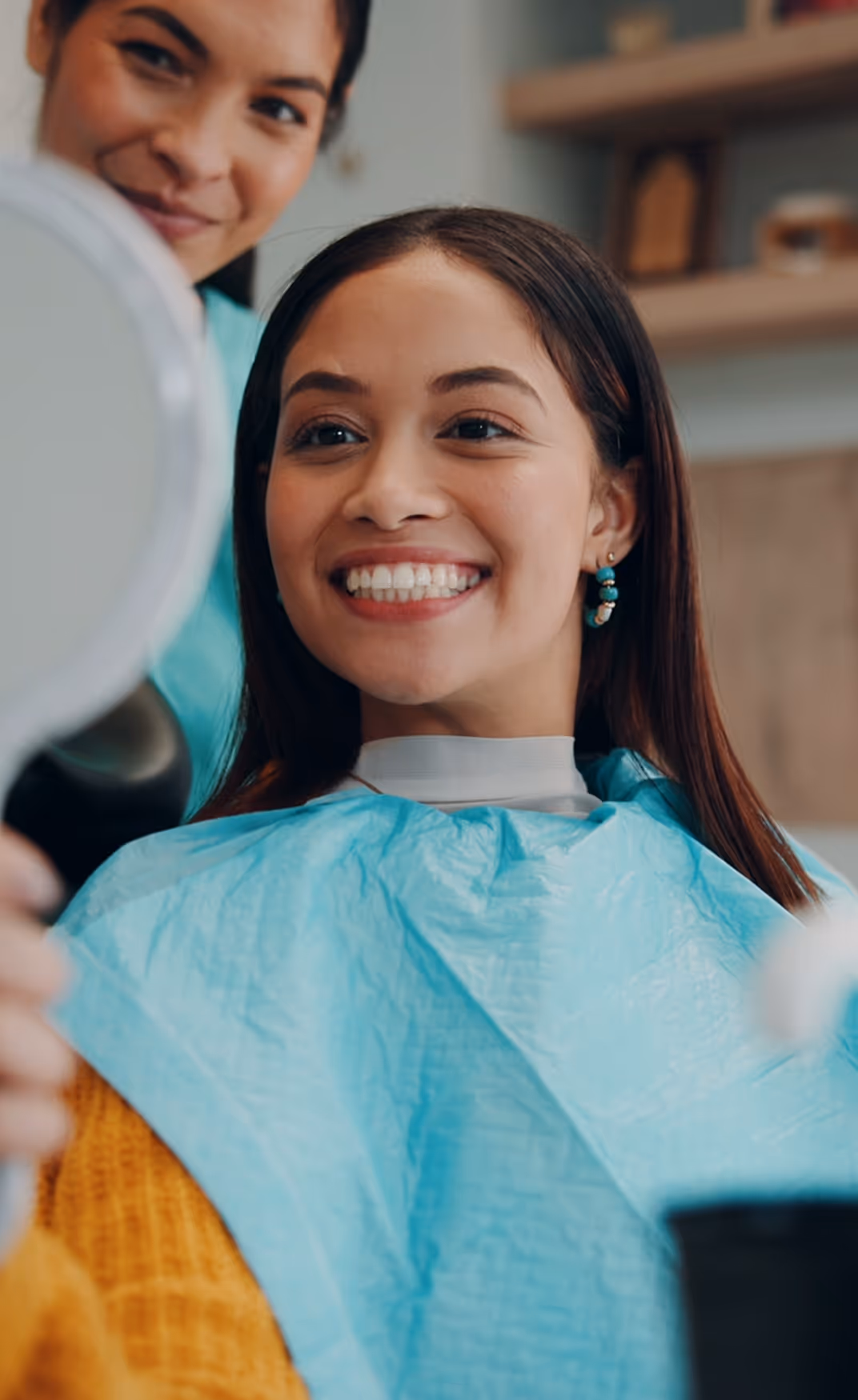 Young woman in a dental bib smiling and looking at herself in a handheld mirror while a dentist watches happily.