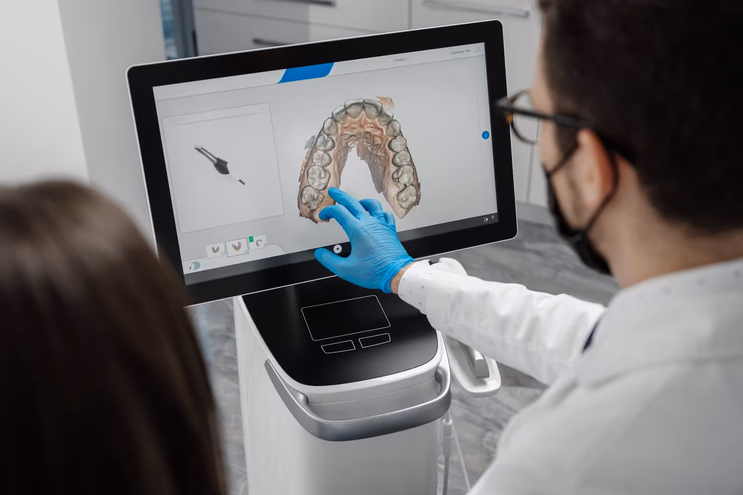 Dentist wearing blue gloves points at a 3D digital dental scan on a touchscreen monitor in a dental office.
