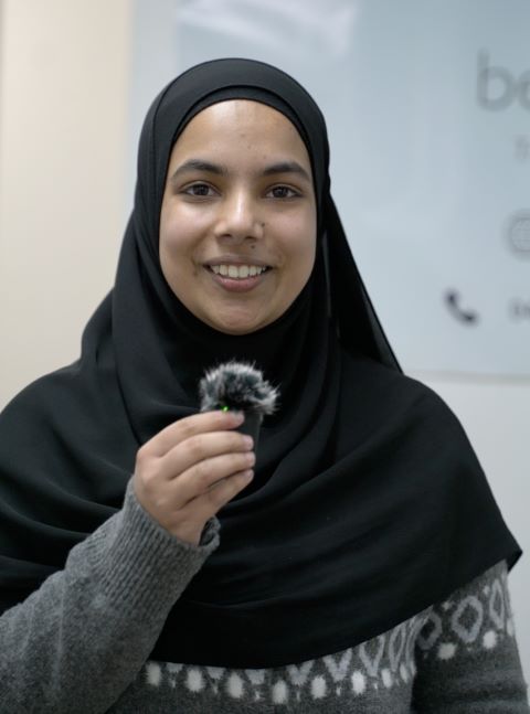 Young woman wearing a black hijab and gray sweater smiling while holding a small microphone with a furry windscreen.