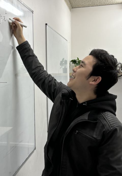 A man in a black jacket writing mathematical equations on a whiteboard with a marker.