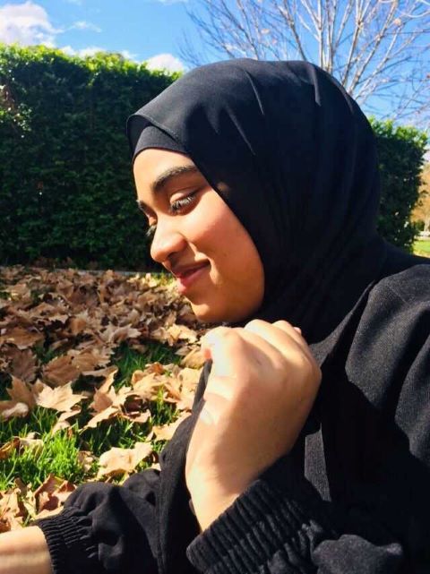 Young woman wearing a black hijab and jacket smiling outdoors with autumn leaves and green hedge in the background.