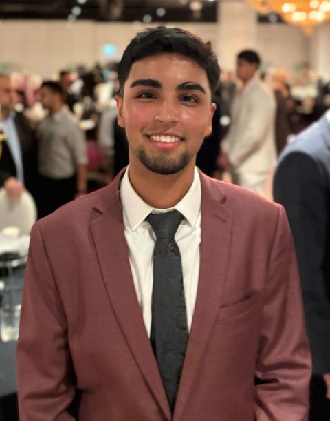 Young man smiling, wearing a maroon suit jacket, white shirt, and dark tie at a formal event.