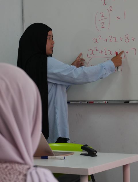 Woman in a black hijab and light blue shirt pointing at a whiteboard with math equations in red marker.