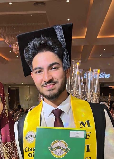Smiling young man wearing a graduation cap and yellow stole holding a green folder at a graduation ceremony.