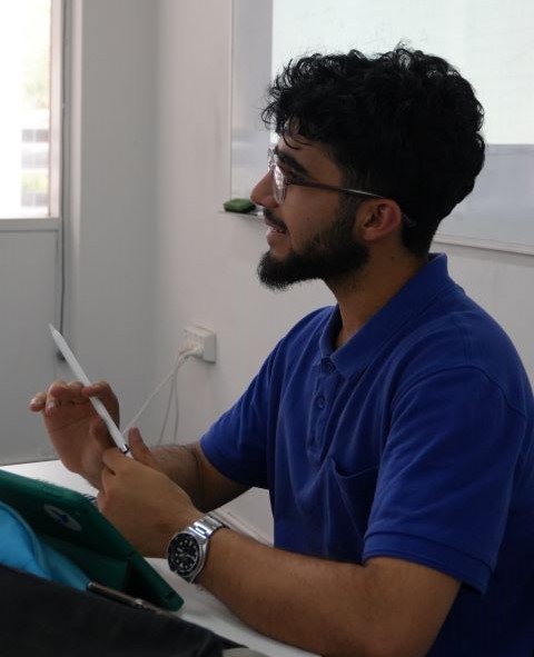Man with glasses and beard wearing a blue polo shirt holding a pen and paper, sitting at a table in a room with a whiteboard.