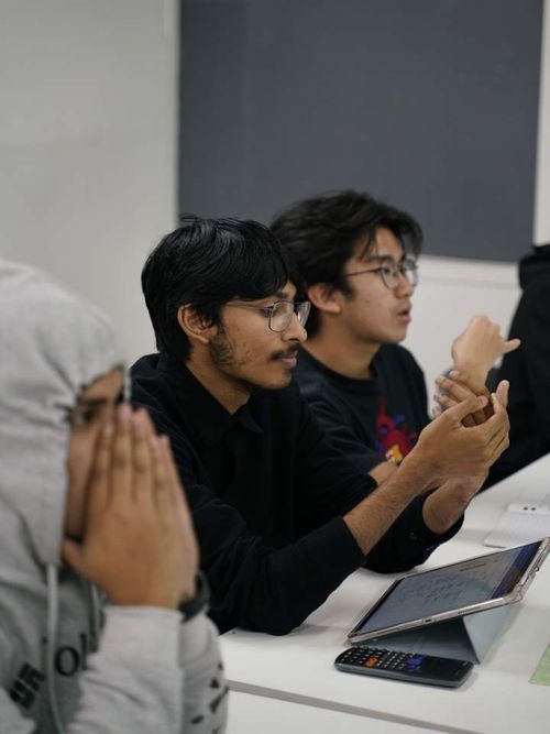 Three young men sitting at a table, two engaged in discussion with an open tablet and calculator in front of them, one person in a gray hoodie with hands near face.