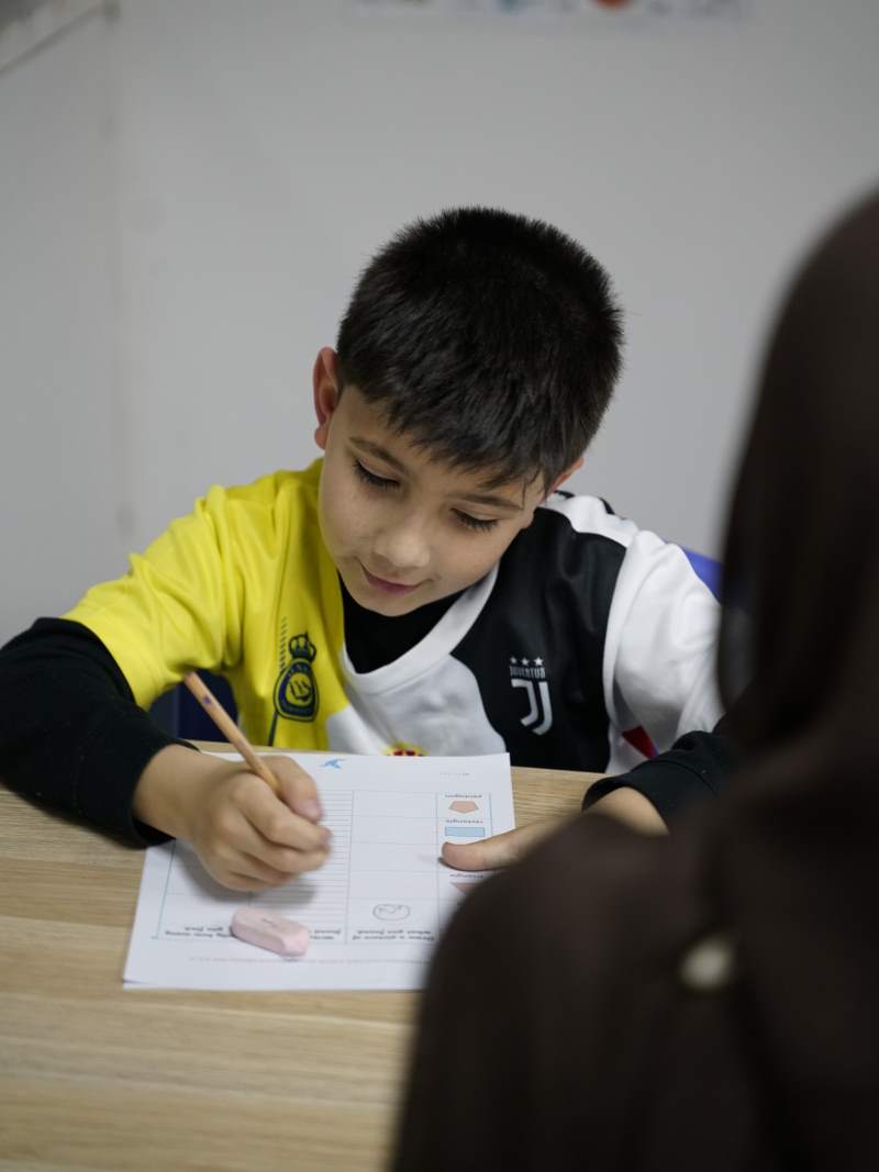Young boy in a black, white, and yellow shirt writing on a worksheet with a pencil at a wooden table.