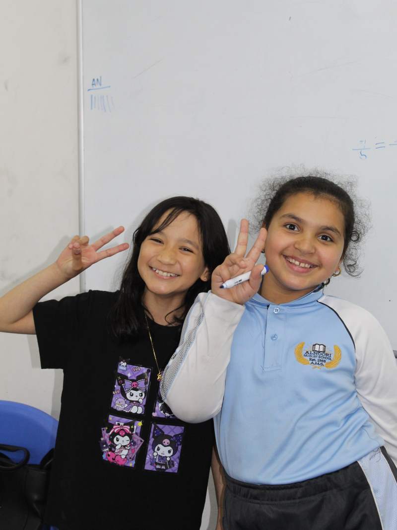 Two smiling girls posing indoors, making peace signs with their hands against a whiteboard background.