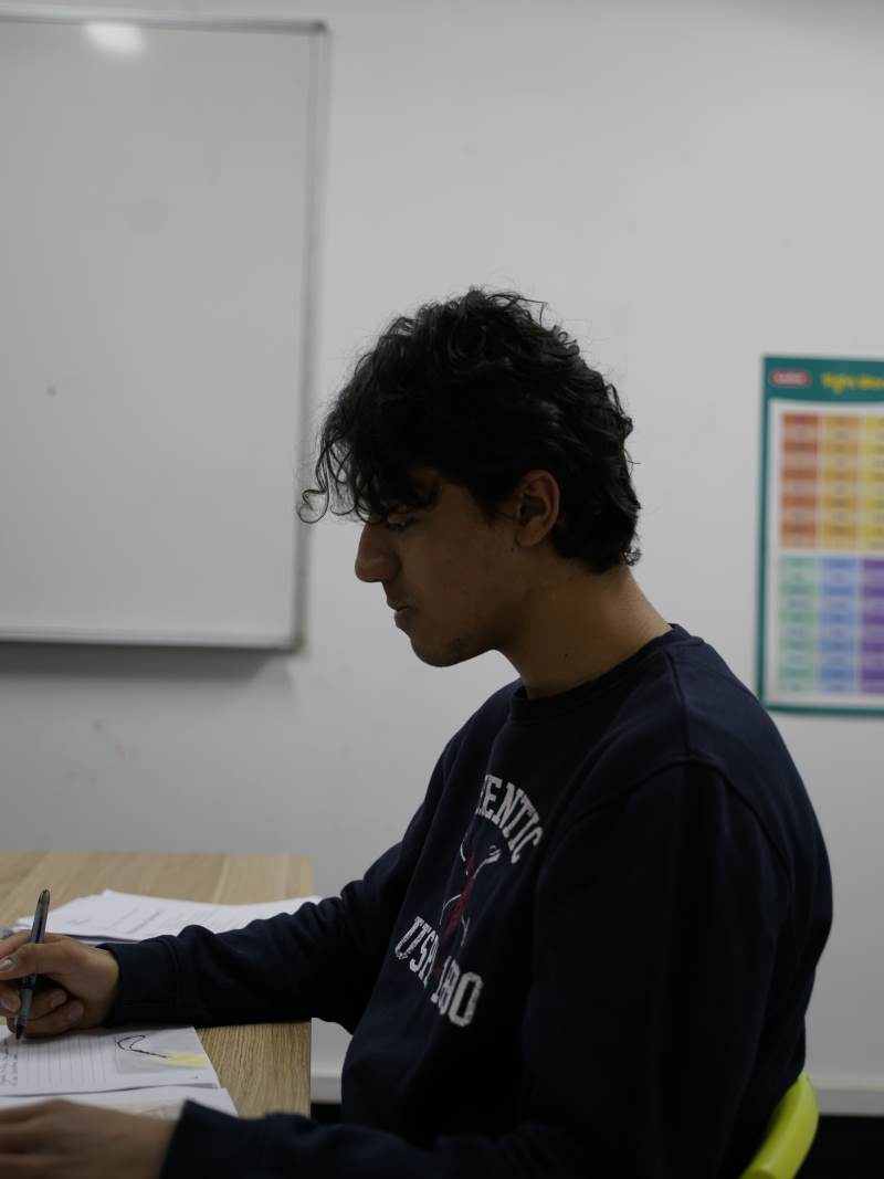 Young man with curly dark hair writing on paper at a wooden table in a classroom with a whiteboard and colorful chart in the background.