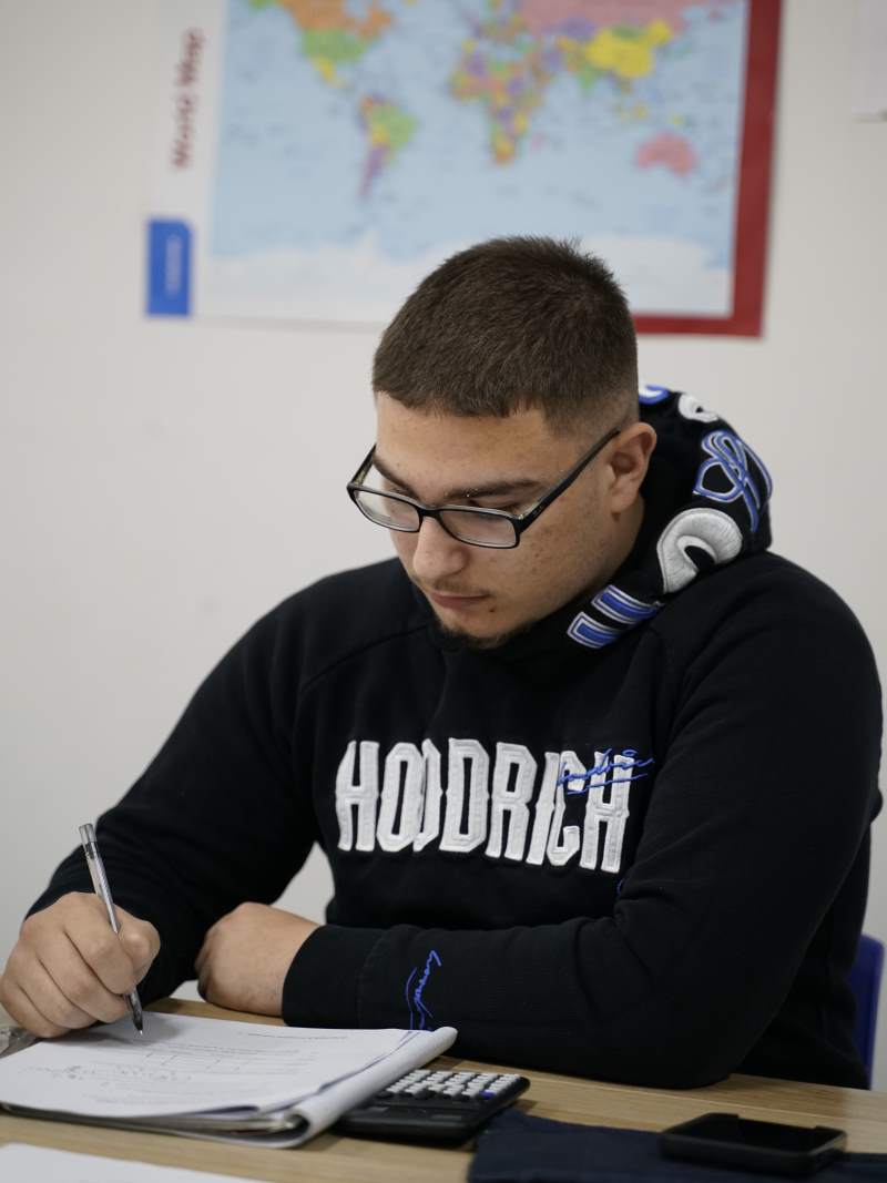 Young man wearing glasses and a black Hoodie is writing on paper with a pen at a desk with a calculator and smartphone, with a world map on the wall behind him.
