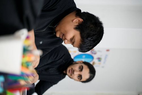Two young men sitting at a table, one smiling and looking down, the other looking at him, with blurred colorful art supplies in the foreground.