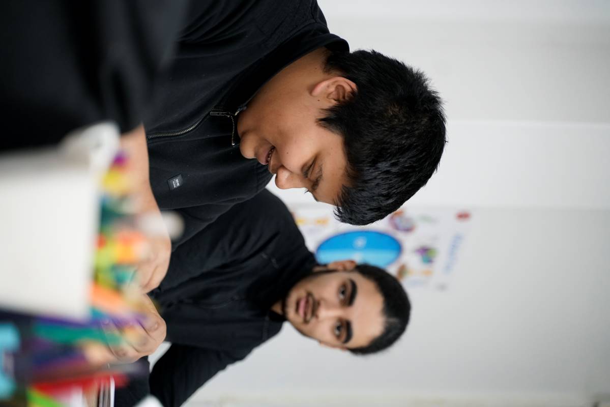 Two young men sitting at a table, one smiling and looking down, the other looking at him, with blurred colorful art supplies in the foreground.