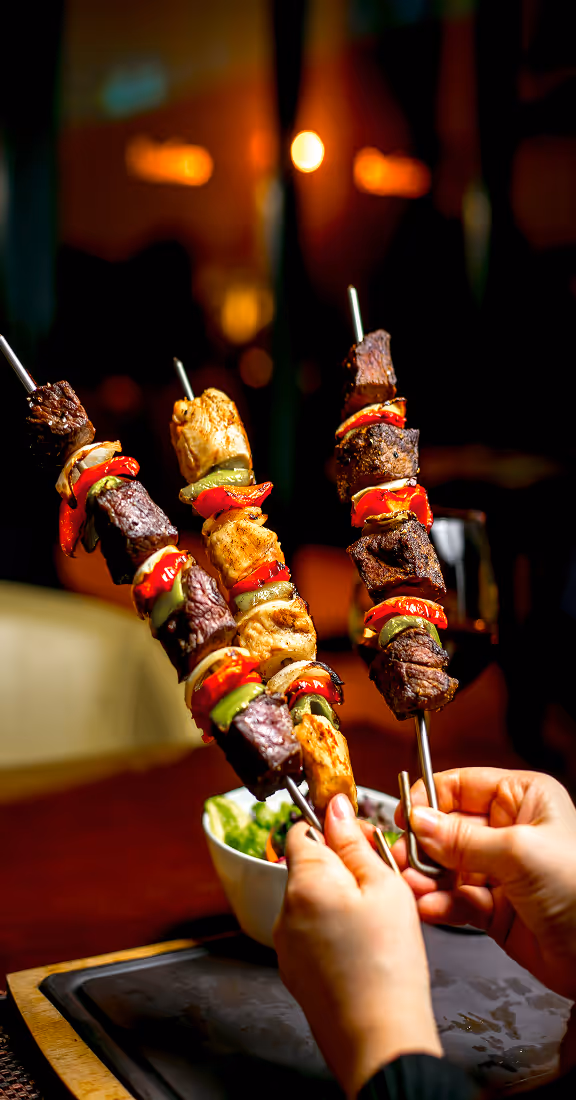 Two hands holding three skewers with grilled beef, chicken, red peppers, and green bell peppers over a dark table setting with blurred warm lights in the background.
