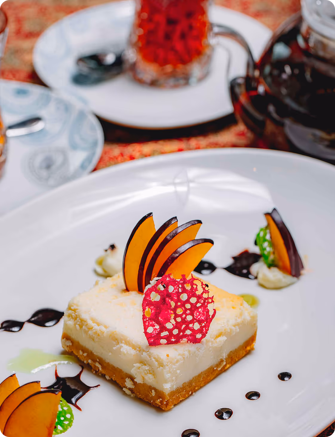 Square slice of cheesecake garnished with thin plum slices and a red decorative crisp on a white plate with artistic sauce dots.