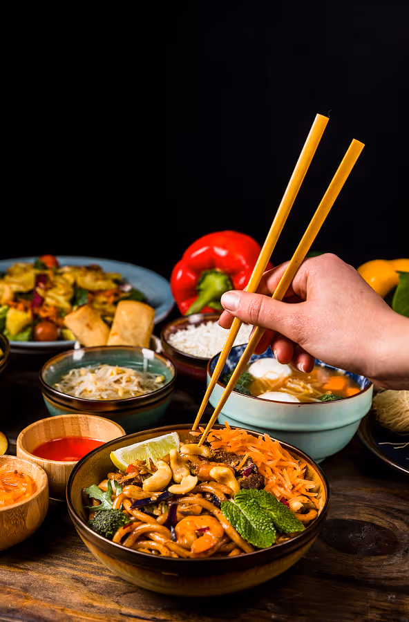Hand using chopsticks to pick up noodles with cashews, lime, and shredded carrots from a bowl surrounded by various Asian dishes and sauces on a wooden table.