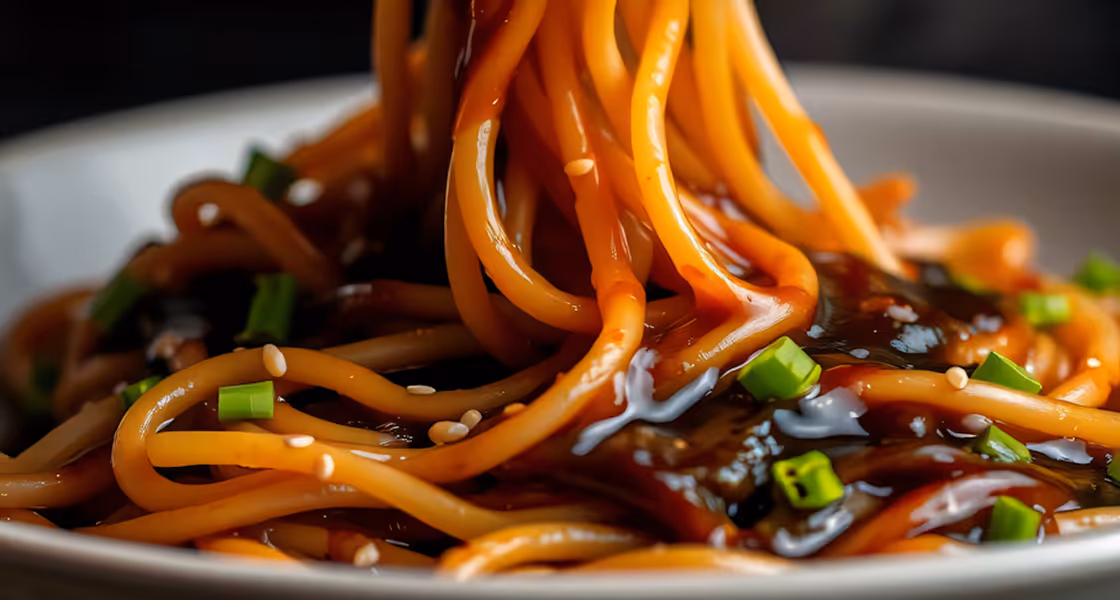 Close-up of noodles coated in dark sauce, garnished with sesame seeds and chopped green onions.