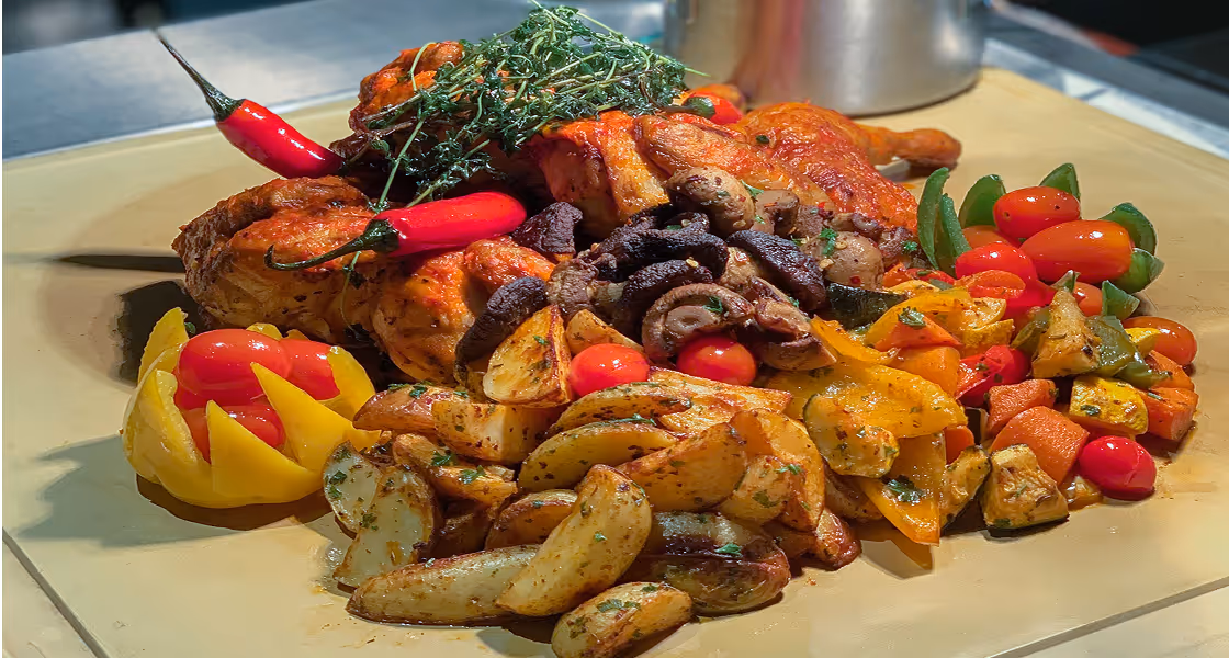 Plate of roasted chicken with herbs, red chili peppers, grilled mushrooms, cherry tomatoes, roasted potato wedges, and mixed grilled vegetables.