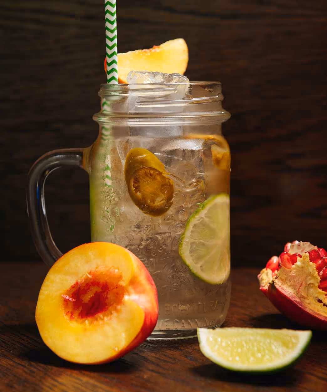 Mason jar filled with iced drink garnished with jalapeño slices, lime slice, and a peach wedge, surrounded by a peach half, lime wedge, and pomegranate pieces on a wooden surface.