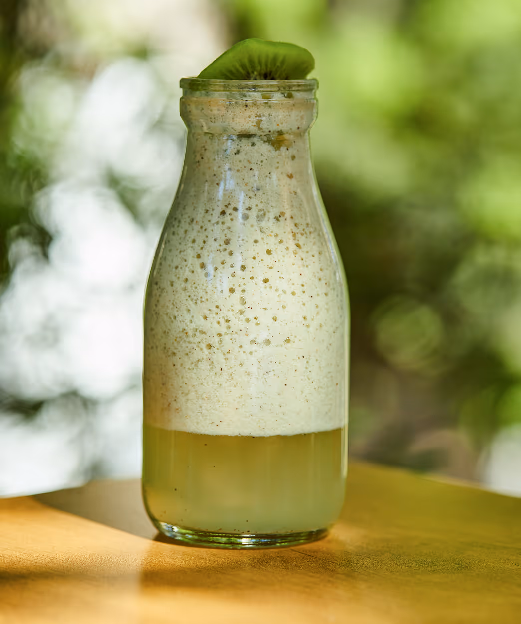 Glass of iced green lemonade garnished with small pink flowers, placed on a wooden board with lemon halves and flower petals scattered around.
