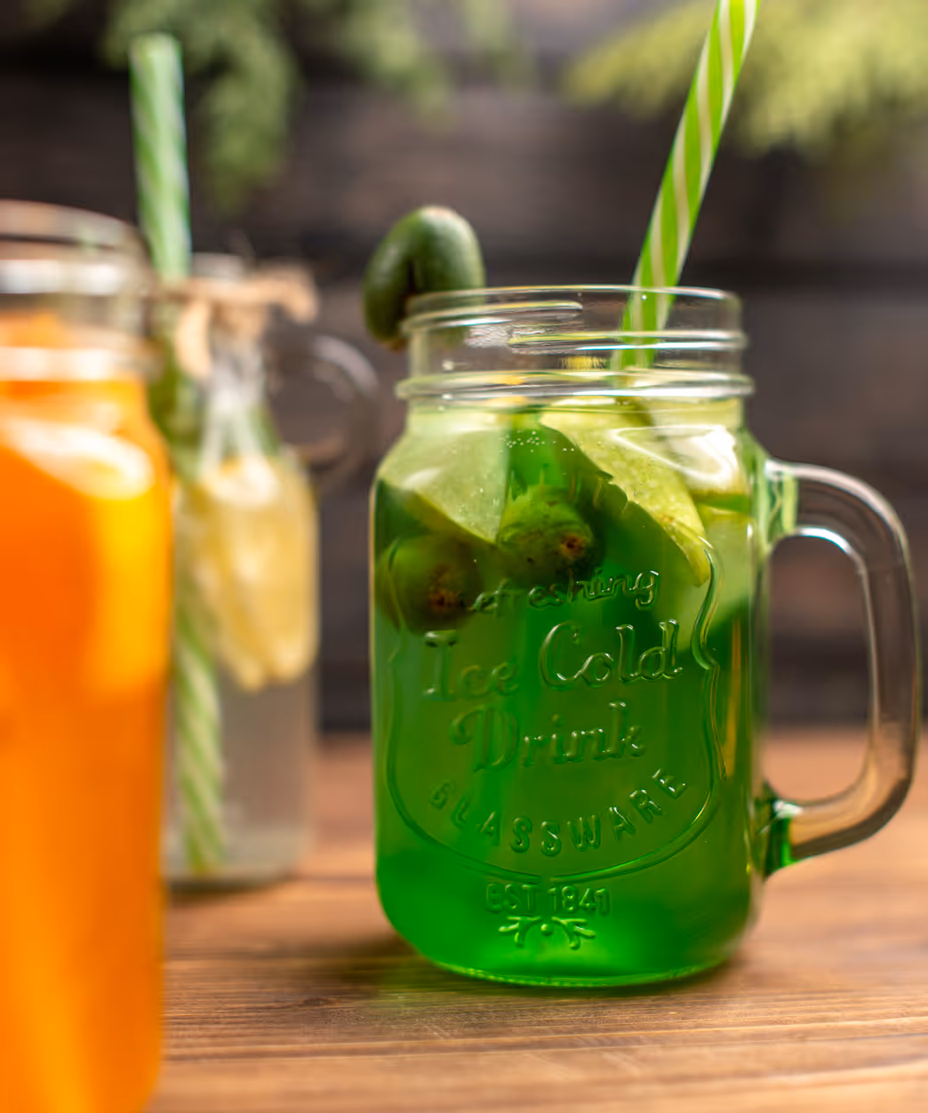 Green drink with lime slices and a striped straw in a glass mason jar on a wooden surface, with blurred orange and clear drinks in the background.