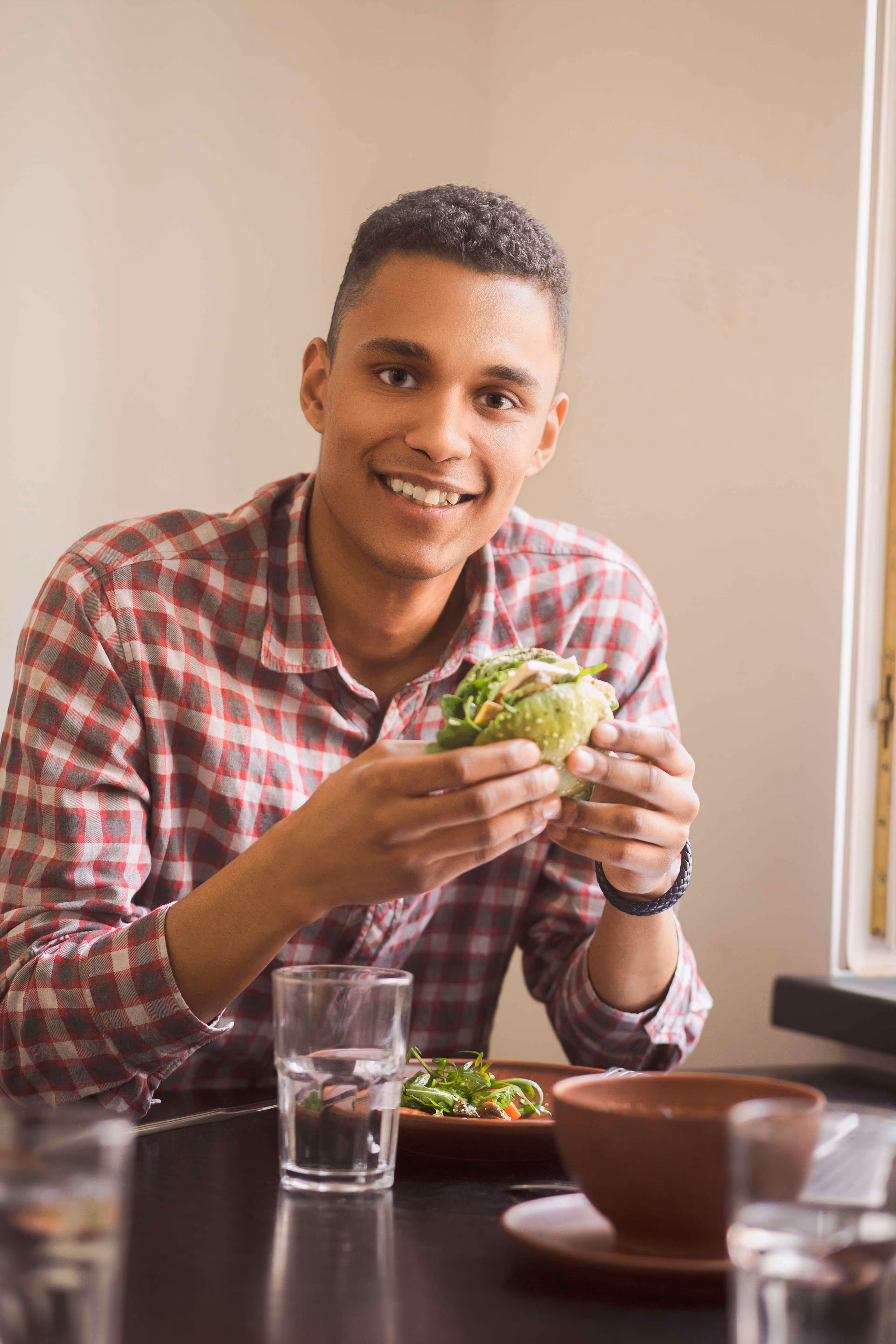 Smiling man in a red plaid shirt holding a sandwich with greens while sitting at a table with a glass of water and a bowl.