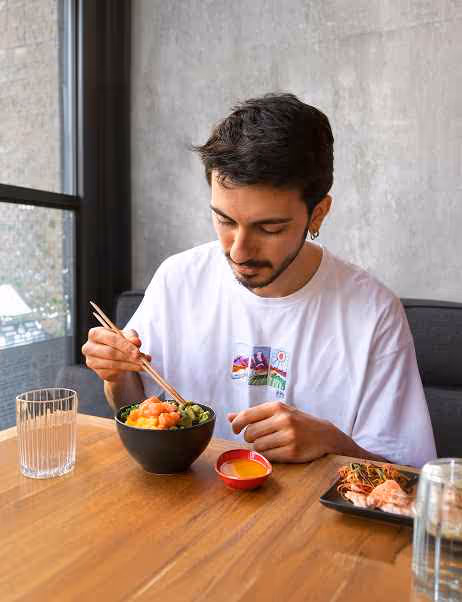 Man wearing a white t-shirt eating a poke bowl with chopsticks at a wooden table near a window.