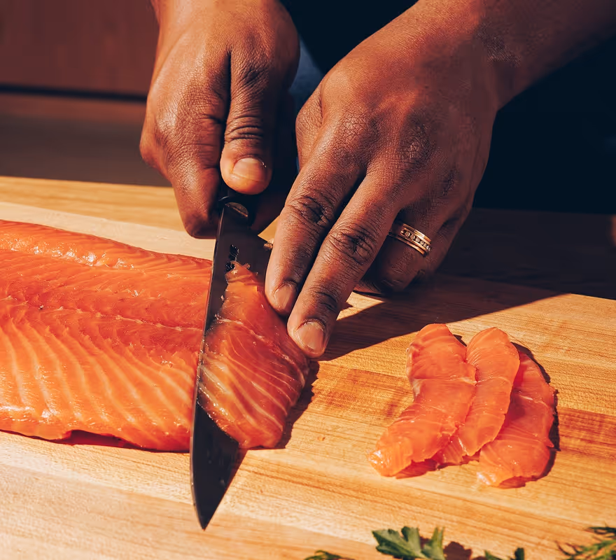 Hands slicing fresh salmon fillet into thin pieces on a wooden cutting board.