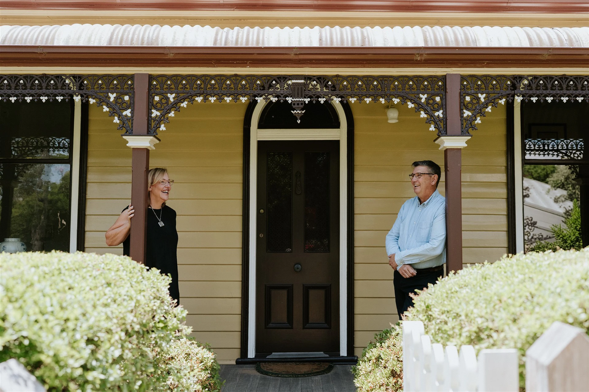 A smiling woman and man stand on opposite sides of a front porch holding wooden posts, facing each other.