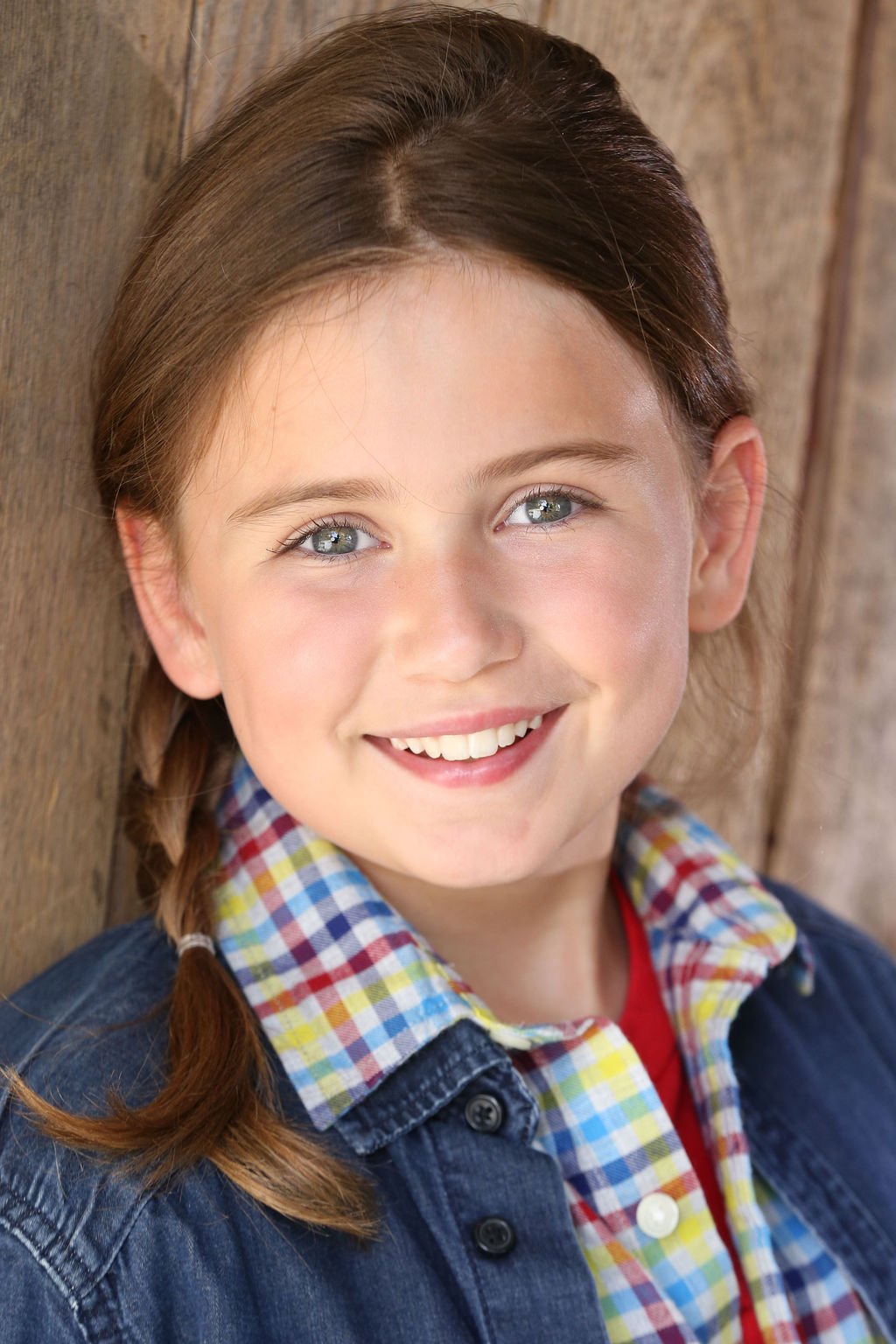 Smiling Caroline Brauner with braided brown hair, wearing a colorful plaid shirt and denim jacket against a wooden background.