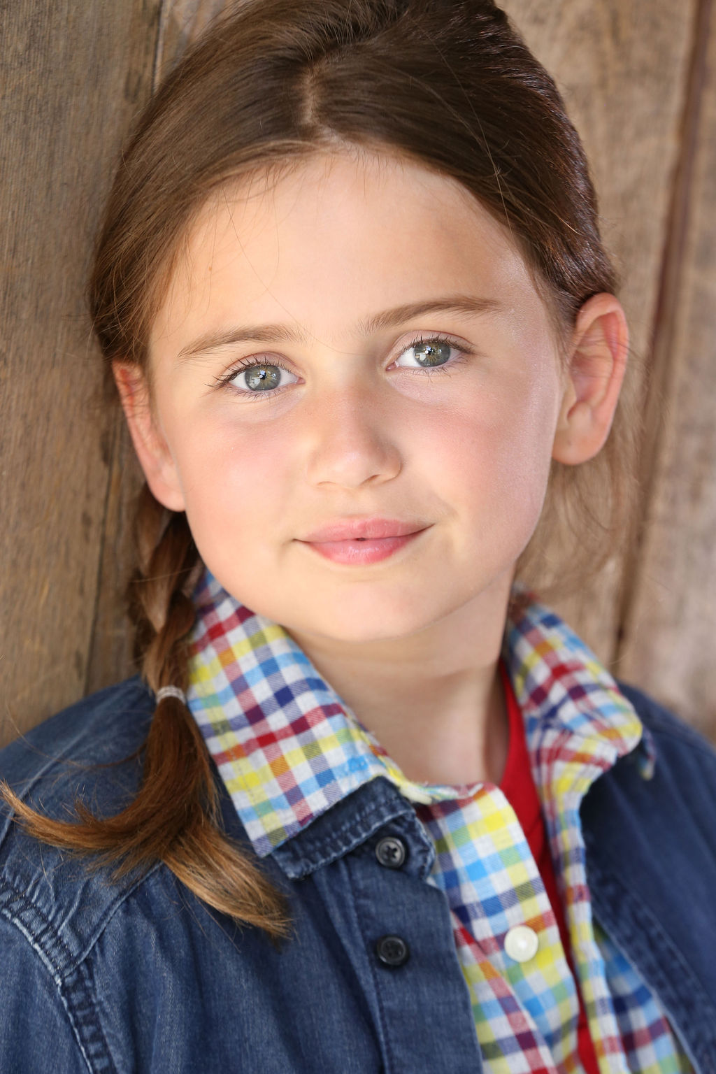 Close-up of Caroline Brauner with light eyes and braided brown hair wearing a blue denim jacket over a multicolored checkered shirt, standing against a wooden background.