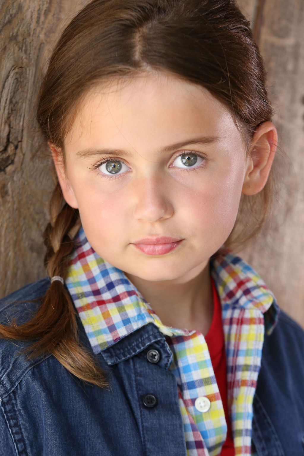 Close-up of Caroline Brauner with light braided hair, wearing a plaid shirt under a denim jacket, standing against a wooden background.