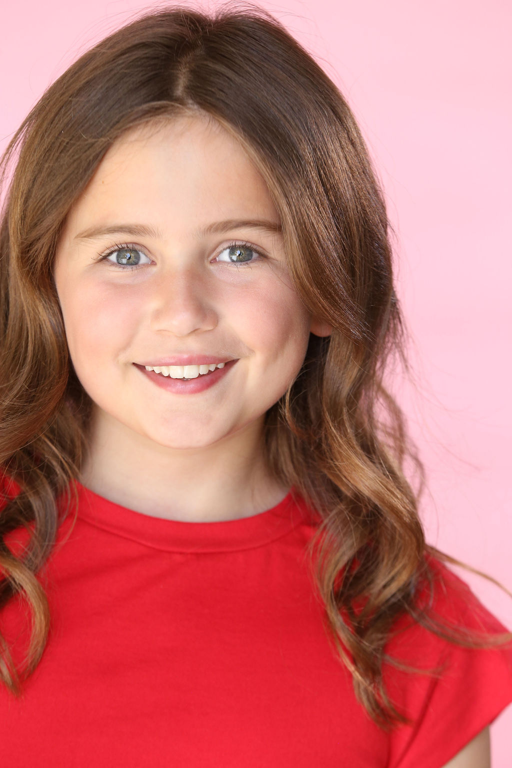Close-up of a smiling Caroline Brauner with long brown hair and blue eyes wearing a red shirt against a pink background.