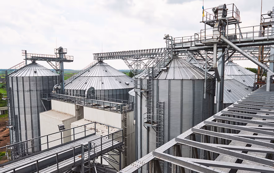 Large metal grain silos and industrial structures under a cloudy sky, viewed from an elevated metal walkway.