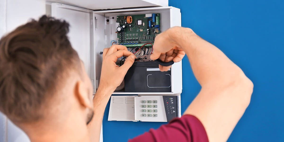 Technician using pliers to work on wiring inside an open security alarm control panel mounted on a blue wall.