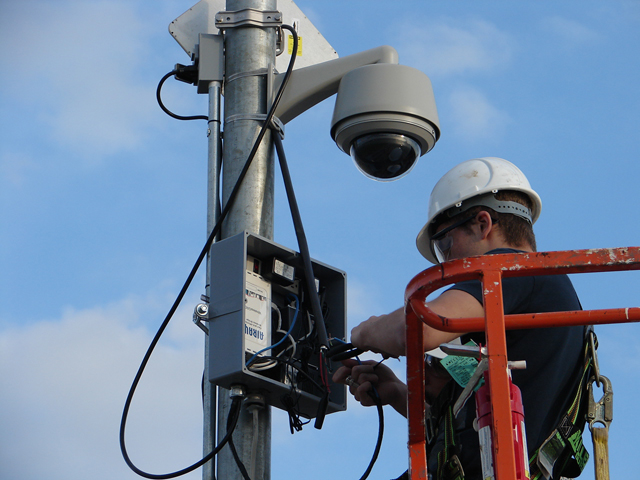 Technician wearing a white hard hat working on electrical wiring inside a metal box mounted on a pole beneath a security camera.