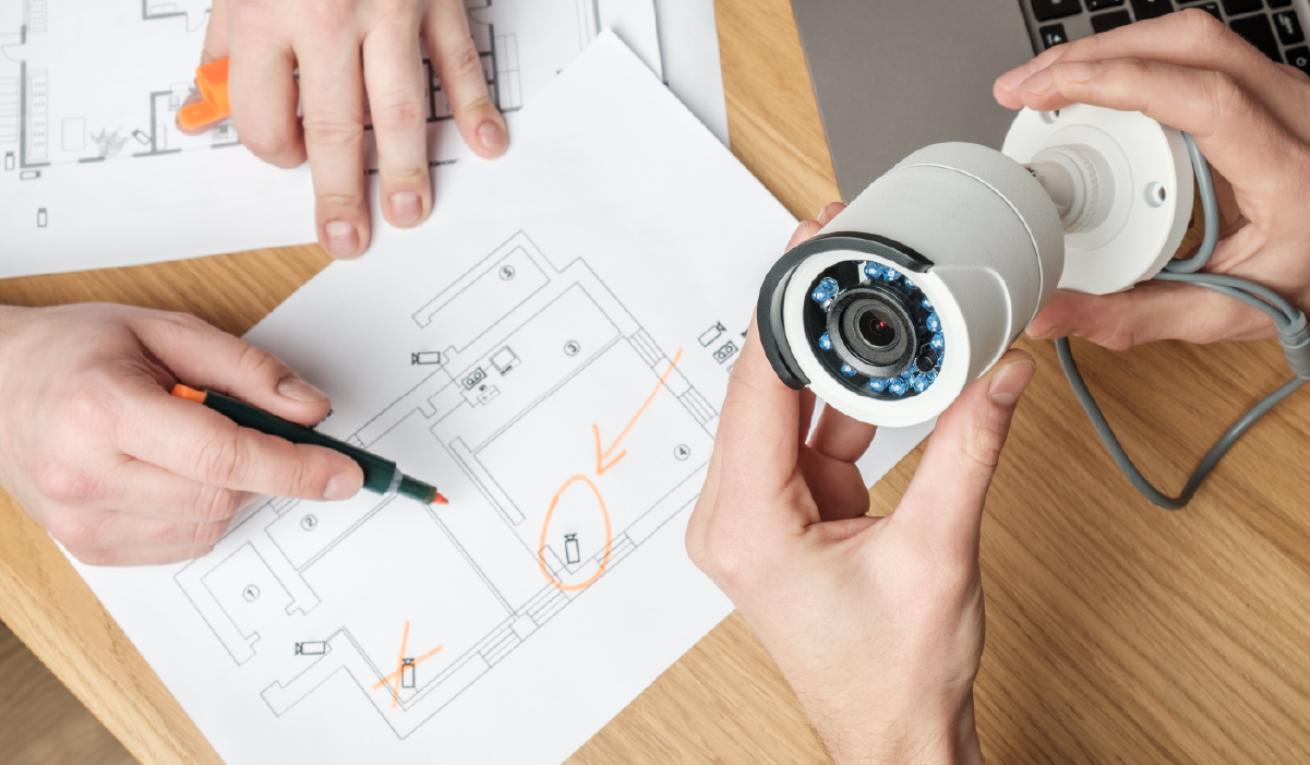 Hands holding a surveillance camera and marking a floor plan with orange pen to indicate camera placement locations.