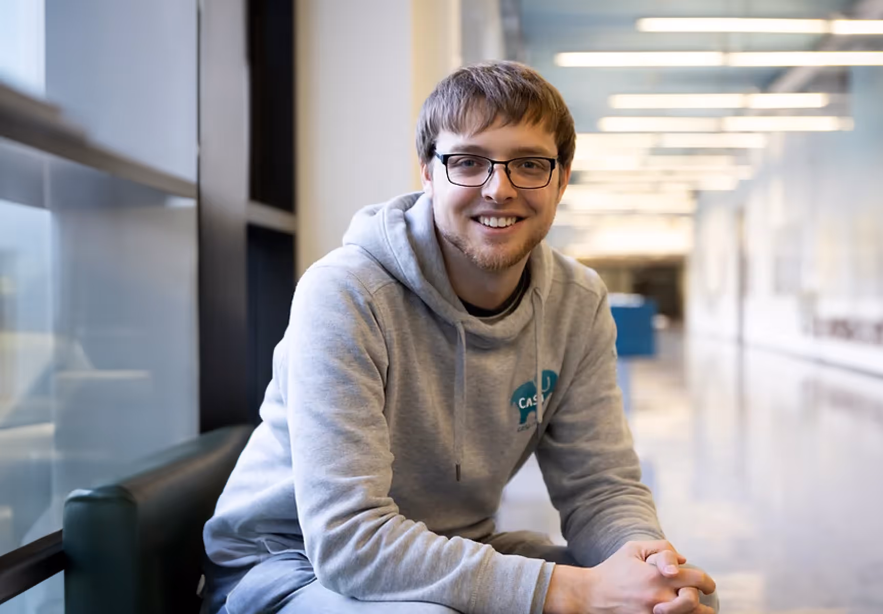 Young man with glasses wearing a light gray hoodie sitting on a chair in a modern, well-lit hallway.
