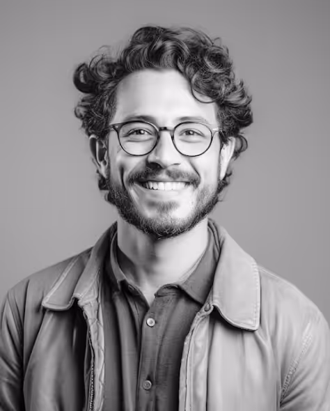 Smiling man with curly hair, beard, and round glasses wearing a collared shirt and jacket in a grayscale portrait.