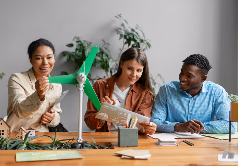Three diverse colleagues discussing a green wind turbine model on a desk with documents and plants.