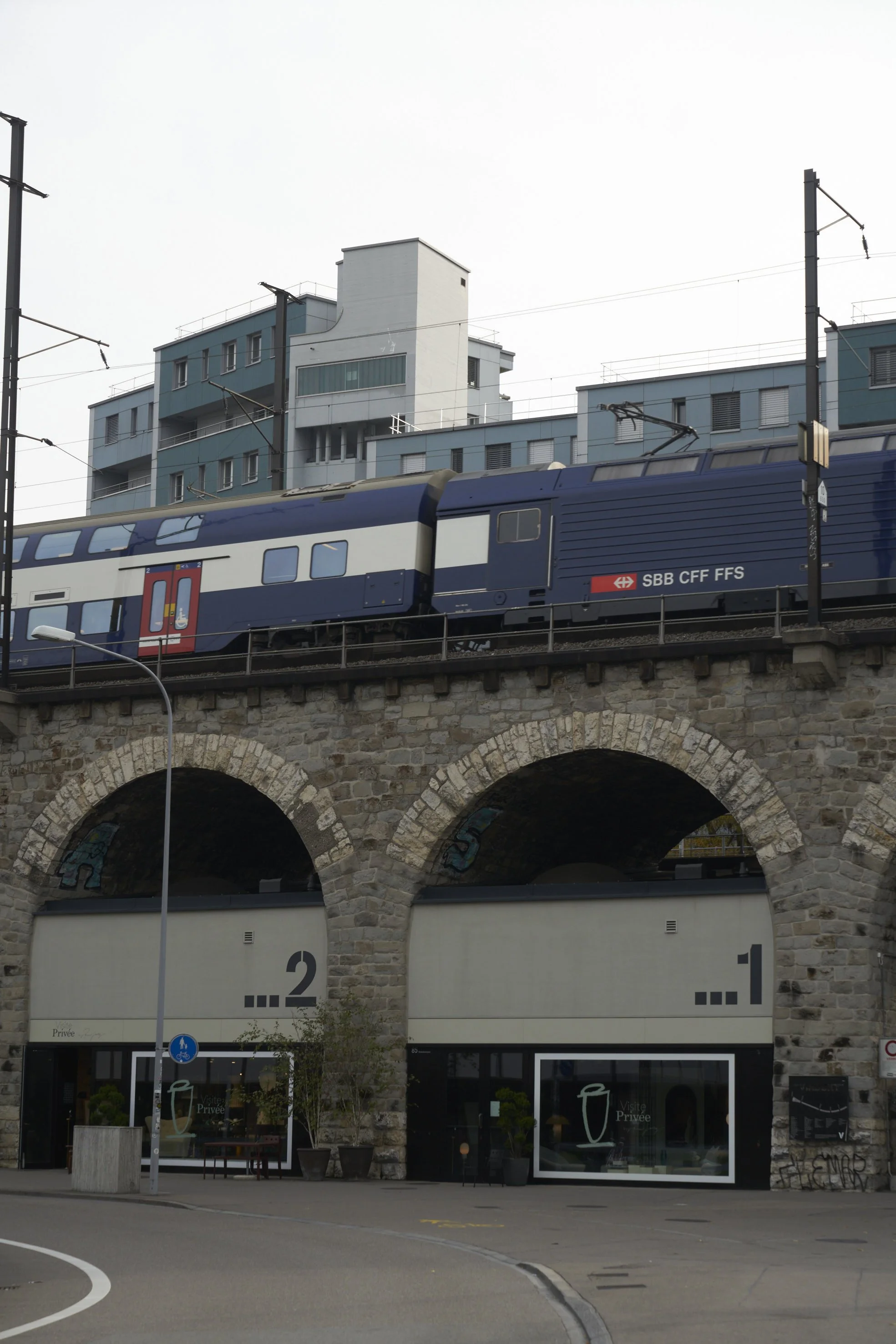 Blue and white SBB CFF FFS train passing on stone arch railway bridge over a street with retail shops below.
