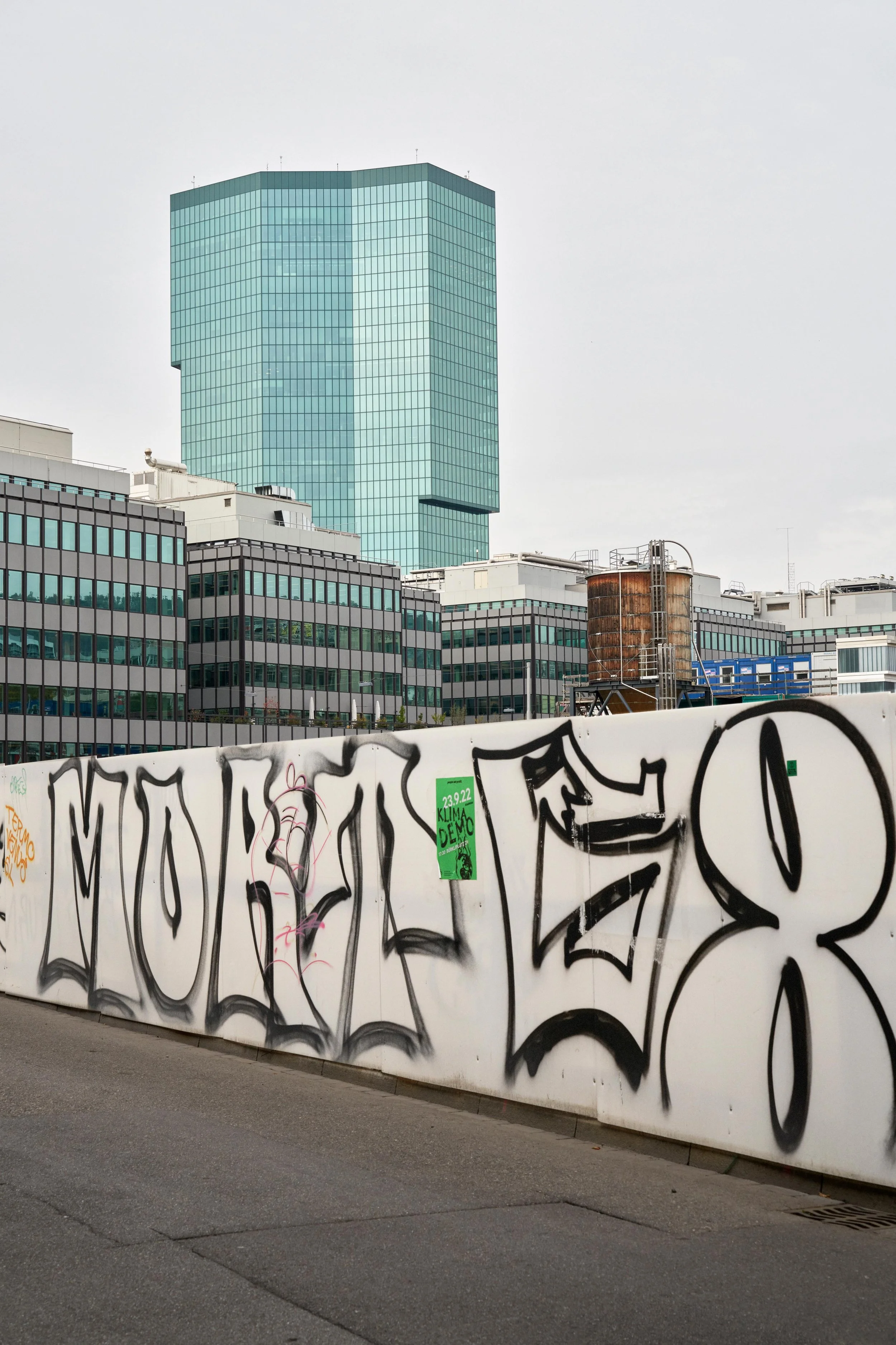 Graffiti spelling 'MORITZ 8' on a white fence with modern office buildings and a tall green glass skyscraper in the background under a cloudy sky.