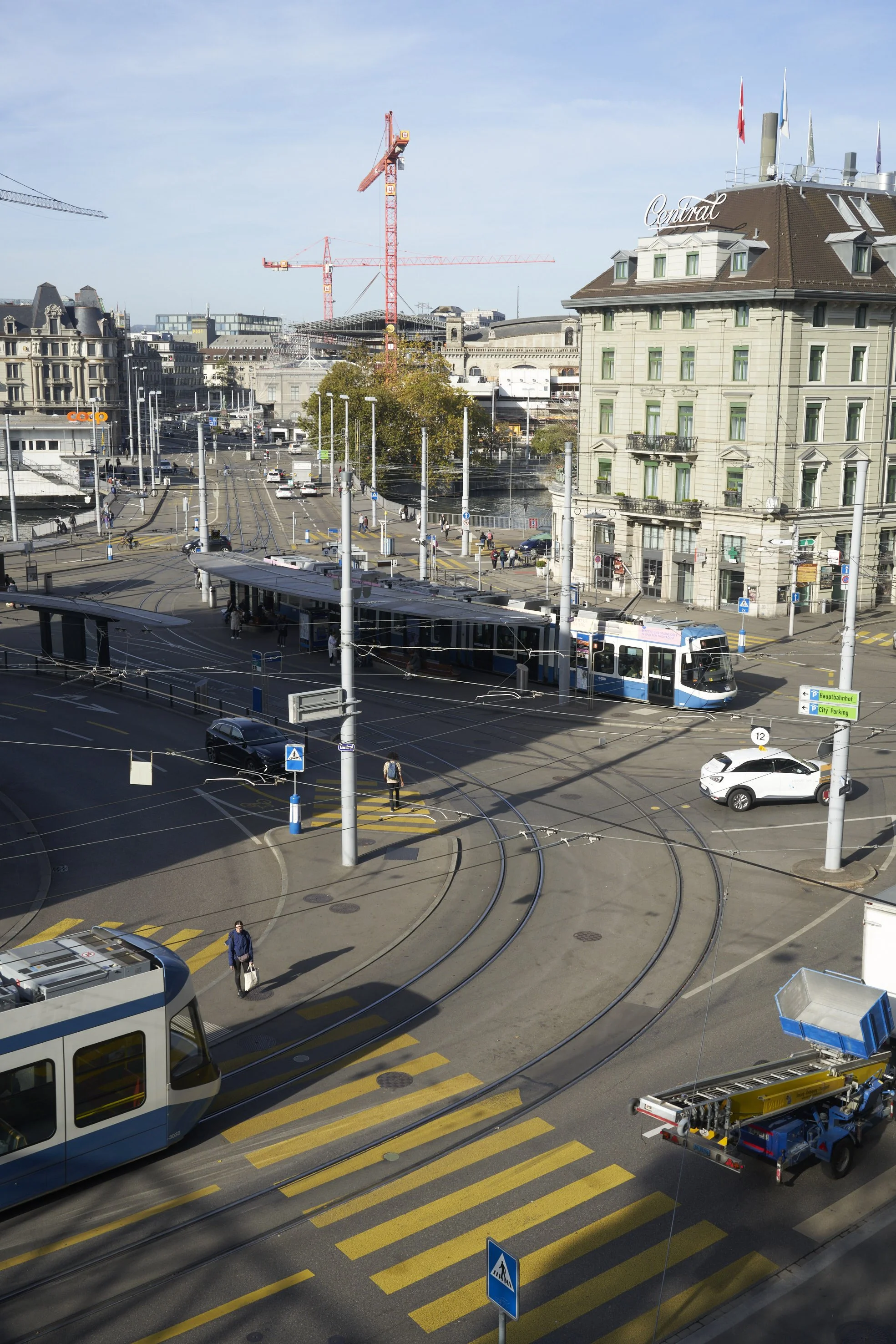 Urban intersection in Zurich with trams, pedestrians, cars, and historic buildings under a clear sky.