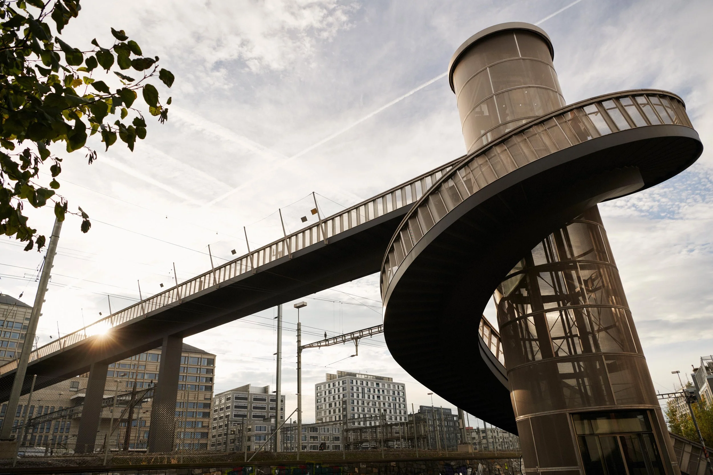 Curved pedestrian bridge with spiral staircase in an urban area with modern buildings at sunset.