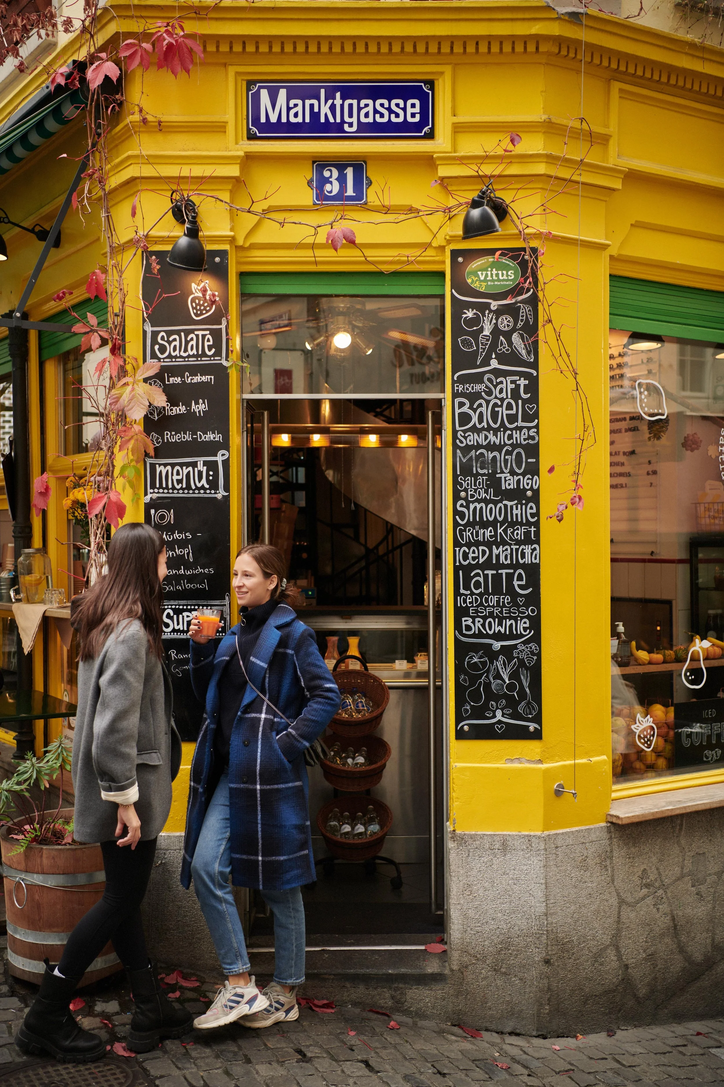 Two women chatting outside a yellow corner café with black chalkboard menus listing salads, bagels, smoothies, and more.