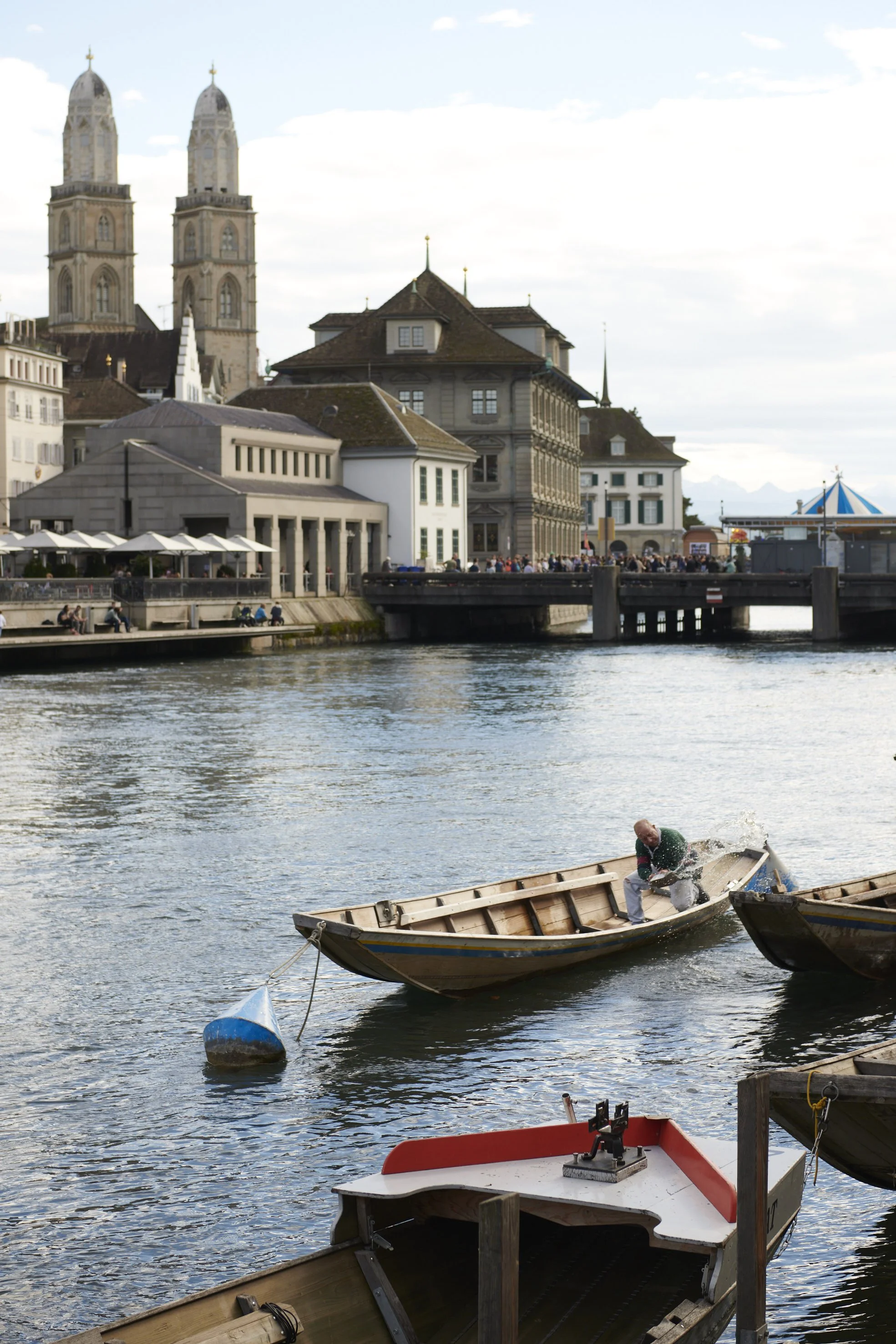 A man splashes water in a wooden boat on a river with historic buildings and a bridge in the background.