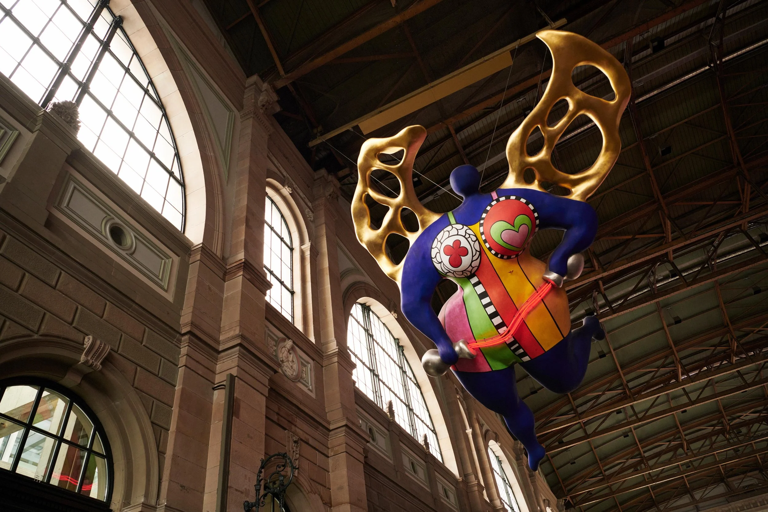 Colorful abstract angel sculpture with golden wings suspended inside a large building with tall arched windows.