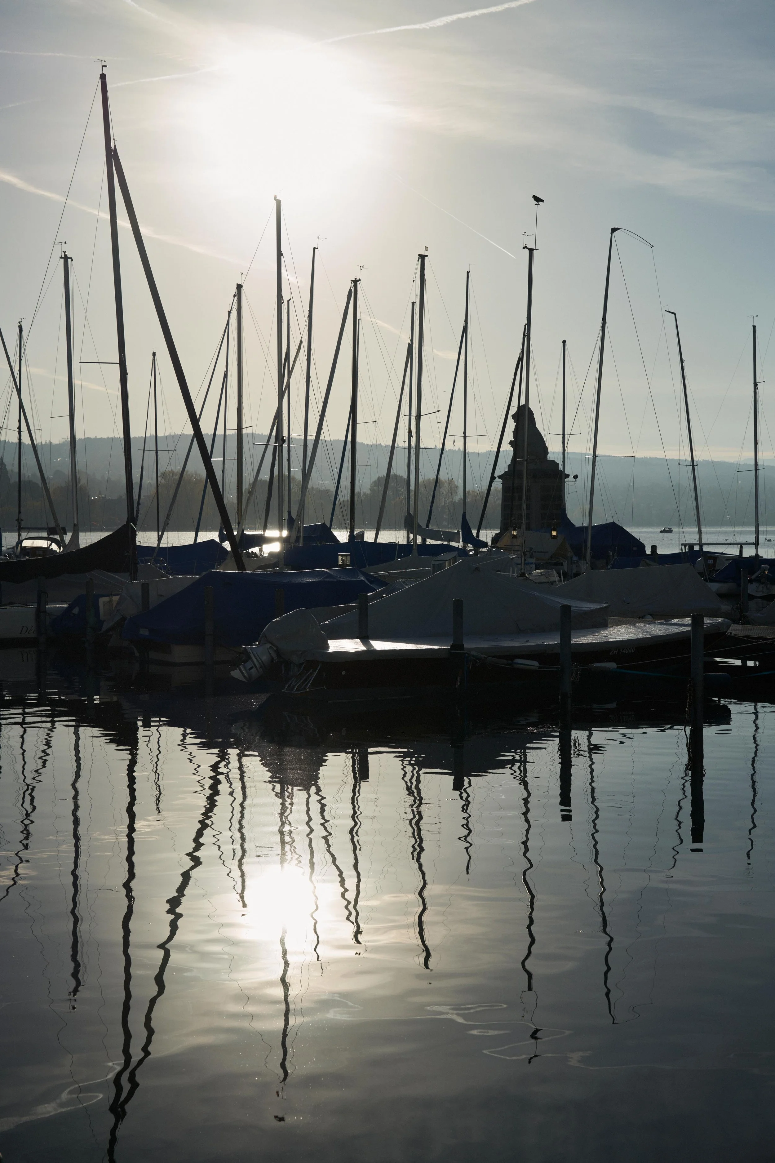Sailboats docked at a marina with masts reflecting in calm water during sunset, with a lion statue visible in the background.