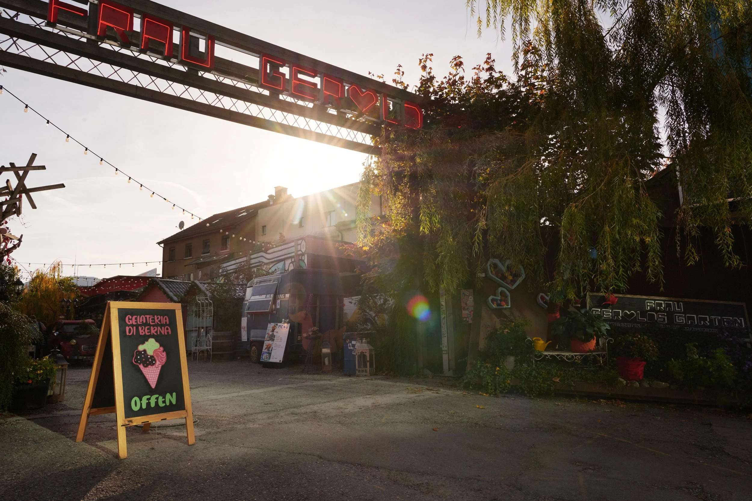 Outdoor view of an ice cream shop with a sign reading 'Gelateria Di Berna Offen' under a sunlit archway with string lights.